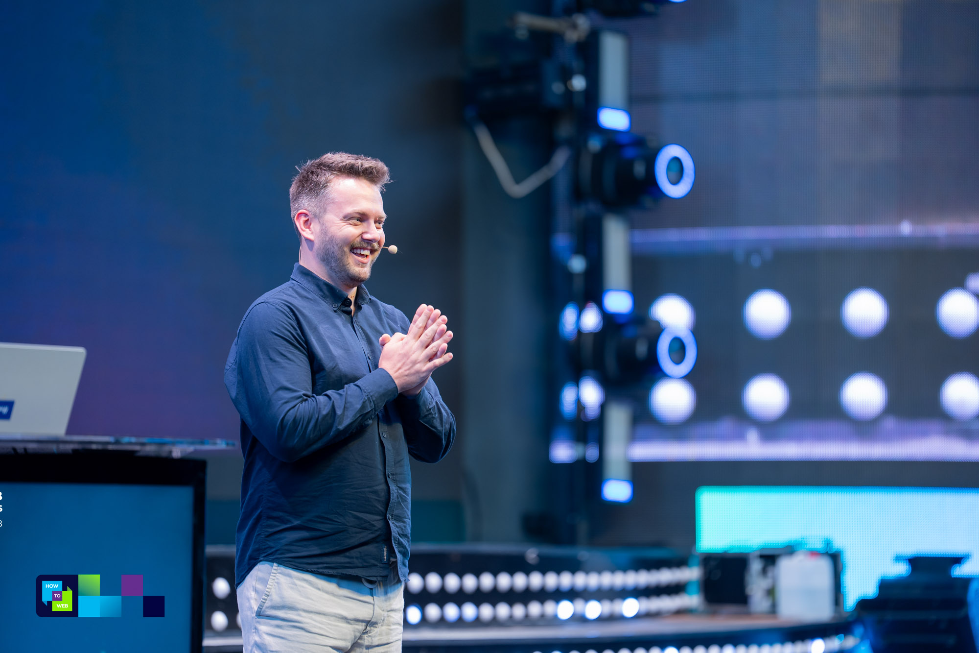 Marcin smiling on the How to Web main stage, hands together, stage lights behind him